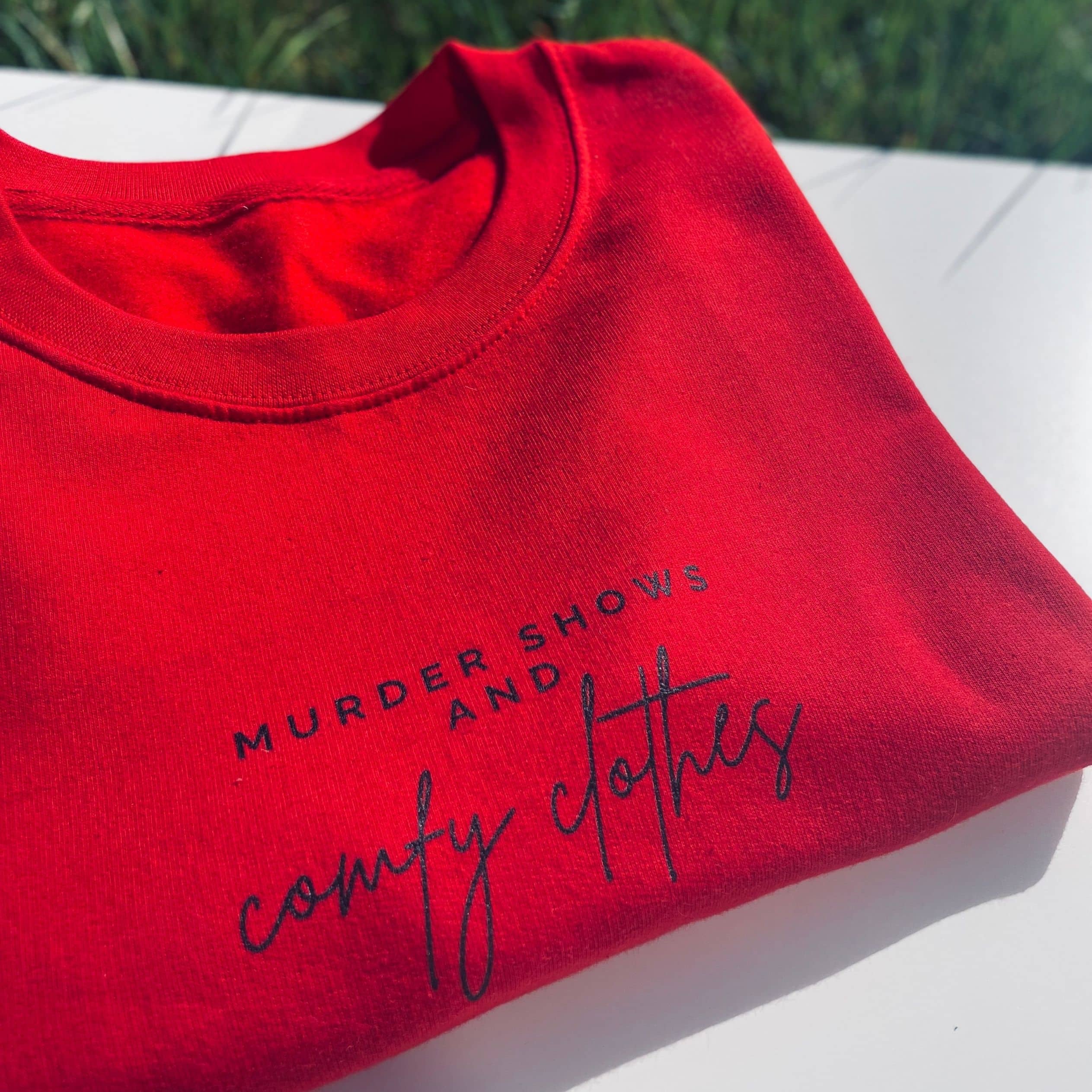 Sweatshirt that says: Murder shows and comfy clothes. Murder shows is in caps and sans serif style font, comfy clothes is in a handwritten style font. Text is small and placed in the centre of the chest, stretching as far as the neckline