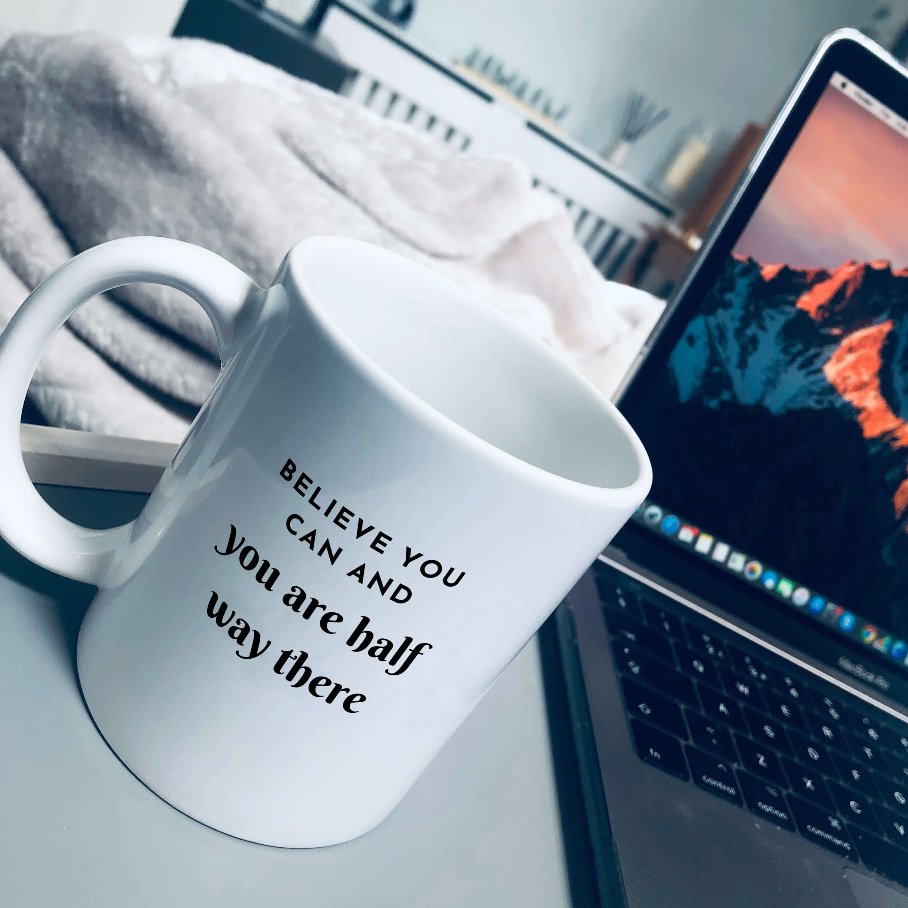Mug on a tray table, with a laptop next to it. Mug reads: believe you can and you are half way there