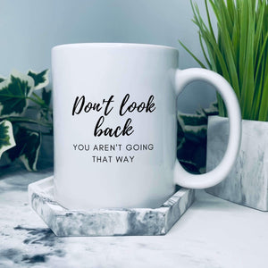 Mug with text in a script style font that is large and says Don't look back. Smaller text underneath in a sans serif style font in caps that says you aren't going that way. Full text reads: Don't look back, you aren't going that way