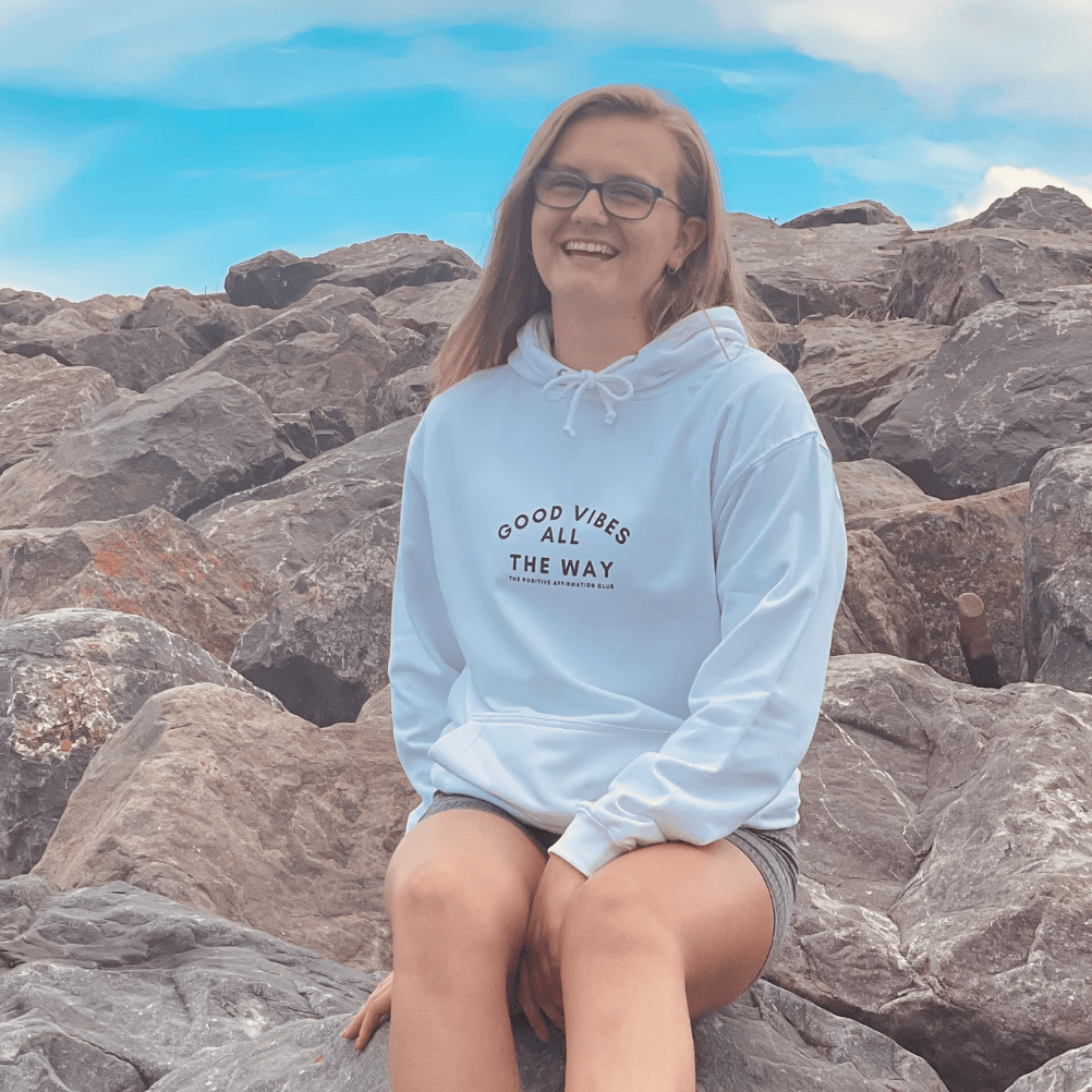 Girl sat on a rocky background with an arctic white good vibes all the way. the positive affirmation club hoody. Hoody that says Good Vibes All The Way. The Positive Affirmation Club. Good Vibes text is curved with alll directly under the middle of good vibes. The way is the directly under that text. In smaller text it then says The positive affirmation club.
