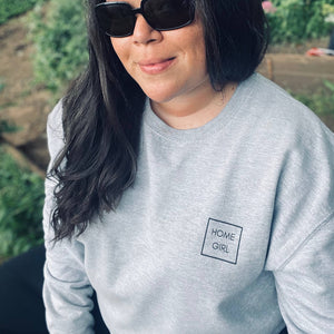 Home Girl in a box in all capitals sans serif font.Heather grey sweatshirt on woman. Woman is in a wooded area with sunglasses on wearing a sweatshirt that says Home Girl on it.  Home Girl is placed top left of the chest